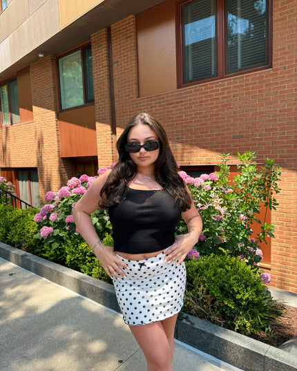 Woman in black top and polka dot skirt standing in front of a brick building with pink flowers.