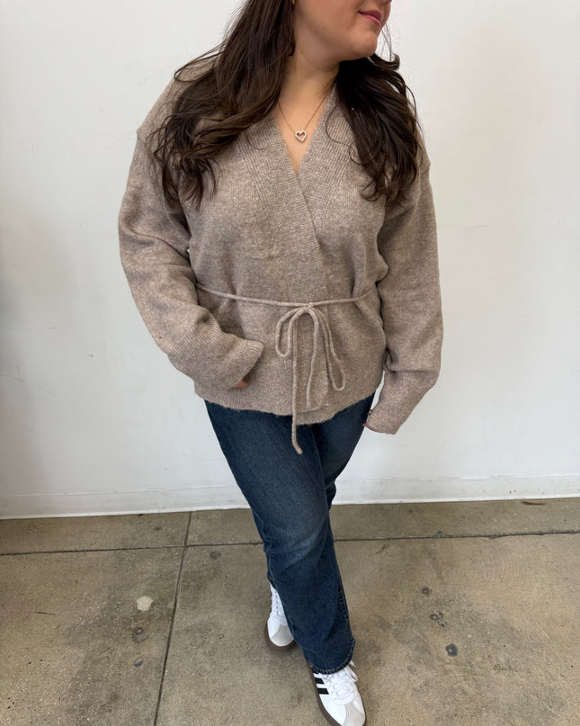 Woman wearing a beige sweater and blue jeans against a white wall.